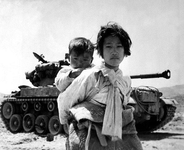 With her brother on her back, a war-weary Korean girl tiredly trudges by a stalled tank, at Haengju, Korea. June 9, 1951. Maj. R.V. Spencer, UAF. (Navy)